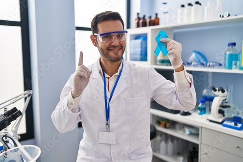 Young hispanic man with beard working at scientist laboratory holding blue ribbon surprised with an idea or question pointing finger with happy face, number one