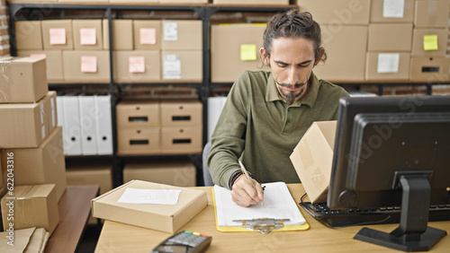 Young hispanic man ecommerce business worker writing on paper checking package at office