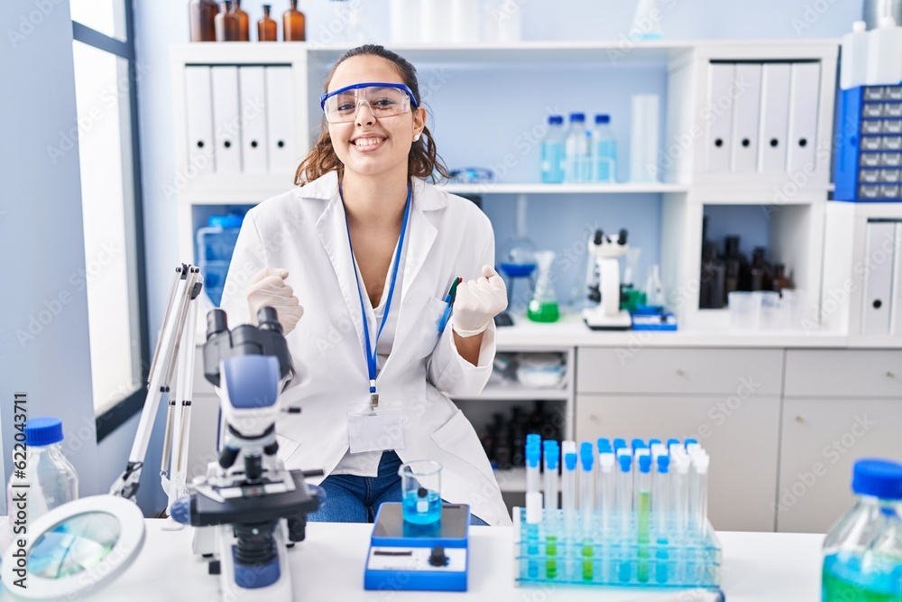 Young hispanic woman working at scientist laboratory very happy and excited doing winner gesture with arms raised, smiling and screaming for success. celebration concept.