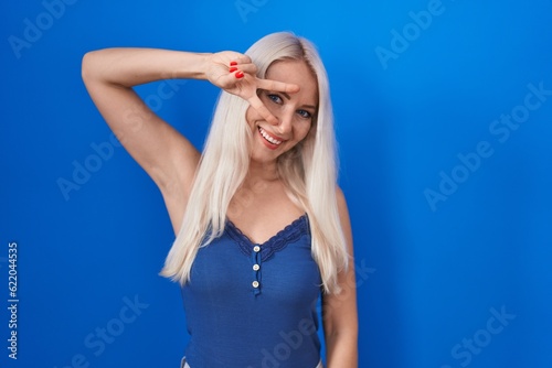 Caucasian woman standing over blue background doing peace symbol with fingers over face, smiling cheerful showing victory © Krakenimages.com