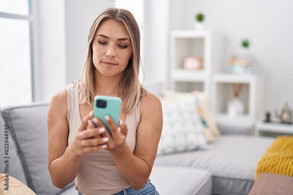 Blonde caucasian woman using smartphone typing message sitting on the sofa thinking attitude and sober expression looking self confident Blonde caucasian woman using smartphone typing message sitting on the sofa thinking attitude and sober expression looking self confident