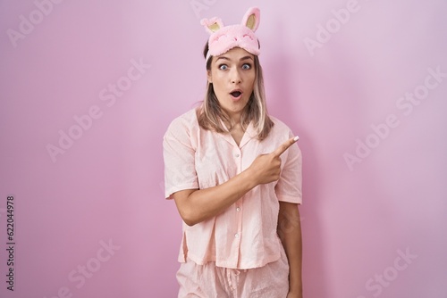 Blonde caucasian woman wearing sleep mask and pajama surprised pointing with finger to the side, open mouth amazed expression.