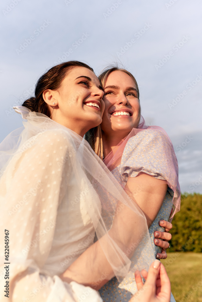 A happy charming bride in a white dress with a white veil and a bridesmaid with a pink veil gently hugging and smiling. Cheerful bachelorette party in the summer park. Close-up portrait. A happy charming bride in a white dress with a white veil and a bridesmaid with a pink veil gently hugging and smiling. Cheerful bachelorette party in the summer park. Close-up portrait.