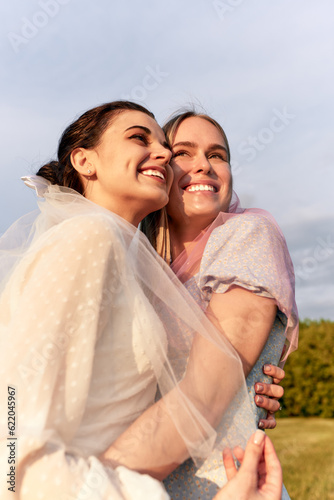 A happy charming bride in a white dress with a white veil and a bridesmaid with a pink veil gently hugging and smiling. Cheerful bachelorette party in the summer park. Close-up portrait.