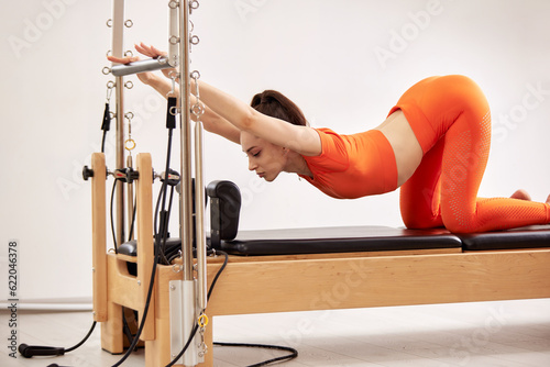 Young girl is doing Pilates on a reformer bed in a bright studio. A slender brunette in an orange bodysuit arches her back and trains body flexibility. Healthy lifestyle concept.