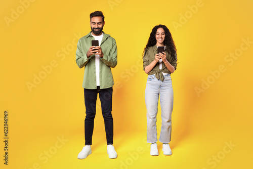 Smiling Middle Eastern Couple Texting Holding Smartphones On Yellow Background