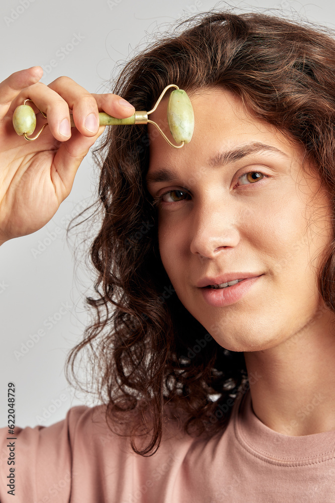 Young handsome long haired guy using jade de-puffing face roller to keep his skin soft and smooth. Caucasian man with brown hair maintaining his natural beauty with skincare routine. Men's self-care. Young handsome long haired guy using jade de-puffing face roller to keep his skin soft and smooth. Caucasian man with brown hair maintaining his natural beauty with skincare routine. Men's self-care.
