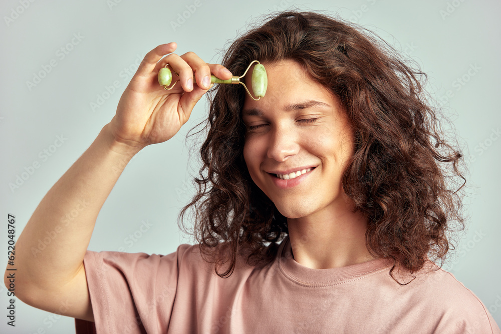 Young handsome long haired guy using jade de-puffing face roller to keep his skin soft and smooth. Caucasian man with brown hair maintaining his natural beauty with skincare routine. Men's self-care. Young handsome long haired guy using jade de-puffing face roller to keep his skin soft and smooth. Caucasian man with brown hair maintaining his natural beauty with skincare routine. Men's self-care.