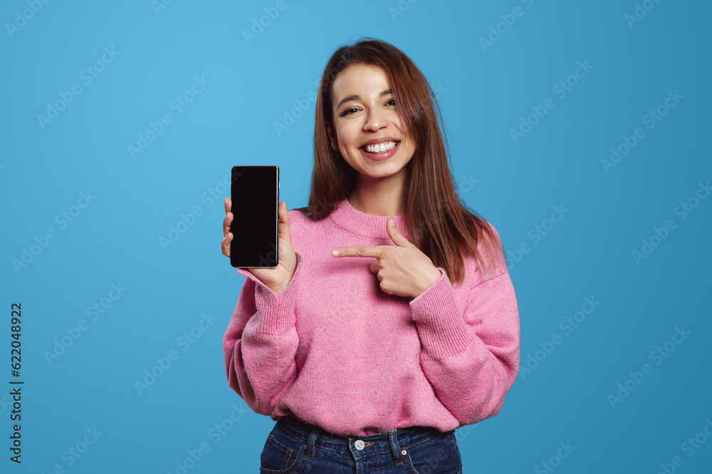 Happy young female in casual pink jumper smiling and holding smartphone while pointing at blank screen, isolated over blue background Happy young female in casual pink jumper smiling and holding smartphone while pointing at blank screen, isolated over blue background
