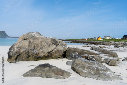de gros rochers sur une plage de sable blanc et des  maisons en fond sous un ciel bleu