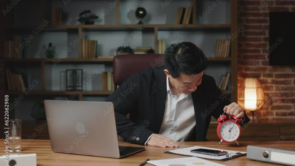 Front view of asian employee sitting at long wooden table with personal computer and cellphone. Happy man in black suit demonstrating red clock meaning that work time is over and leaving office. Front view of asian employee sitting at long wooden table with personal computer and cellphone. Happy man in black suit demonstrating red clock meaning that work time is over and leaving office.