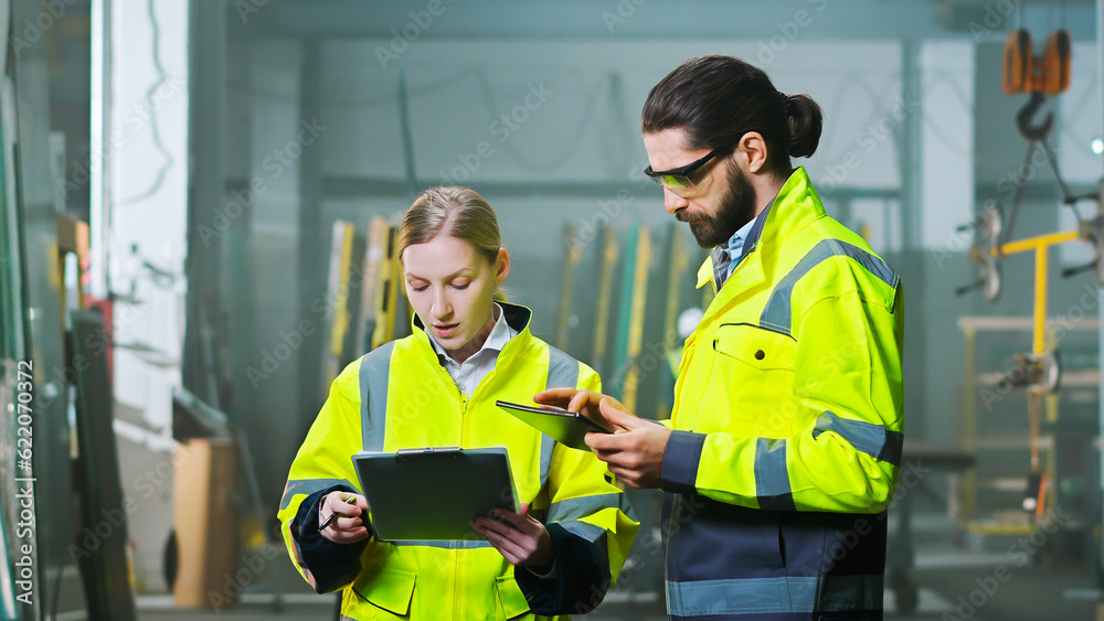 Obraz premium Caucasian man and woman colleagues at manufactory talking and discussing work with tablet device and documents or plan. Production plant. Indoors. Male and female co-workers discussion.