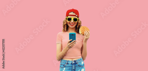 Portrait of stylish smiling young woman with phone and burger fast food on pink background