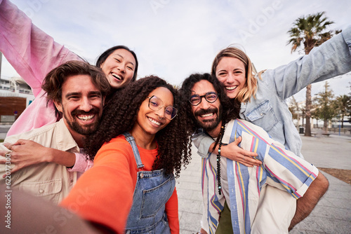 Cheerful group of five friends having fun outdoors, doing piggyback while taking a selfie for social media. Interracial young people enjoying summer, looking at camera happily.