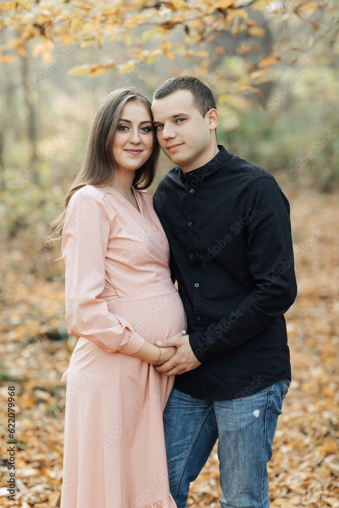 portrait of a happy young family, husband and pregnant wife, standing against the background of beautiful light in the autumn forest, looking at each other portrait of a happy young family, husband and pregnant wife, standing against the background of beautiful light in the autumn forest, looking at each other