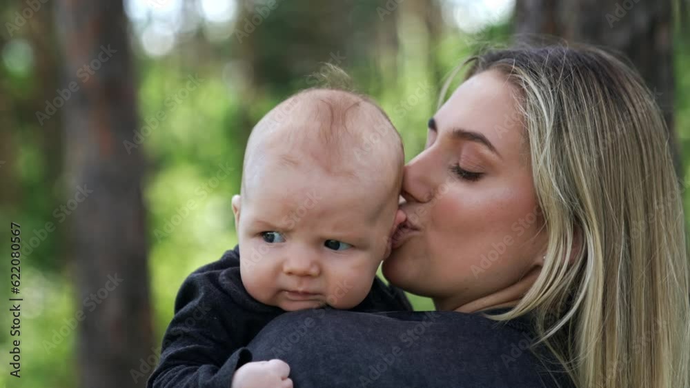 Blonde Caucasian woman holding her baby. Loving mother kissing her child's tiny ear. Close up. Nature at backdrop. Blonde Caucasian woman holding her baby. Loving mother kissing her child's tiny ear. Close up. Nature at backdrop.