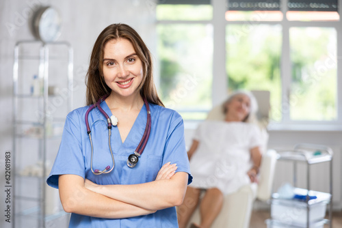 Portrait of friendly young female doctor with stethoscope, wearing uniform standing in treatment room at beauty clinic