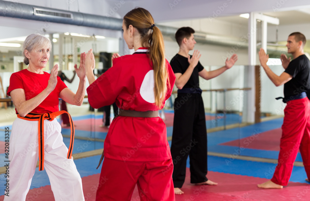 Portrait of concentrated senior woman wearing traditional sportswear sparring with young female opponent during martial arts training in gym