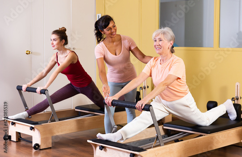 Focused positive elderly woman practicing pilates system on reformer to improve and maintain mobility under supervision of qualified Hispanic female trainer