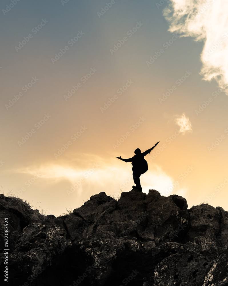 Hiker with arms raised on a mountain facing beautiful sunset silhouette against cloudy sky.