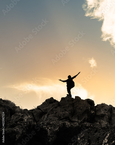 Hiker with arms raised on a mountain facing beautiful sunset silhouette against cloudy sky.