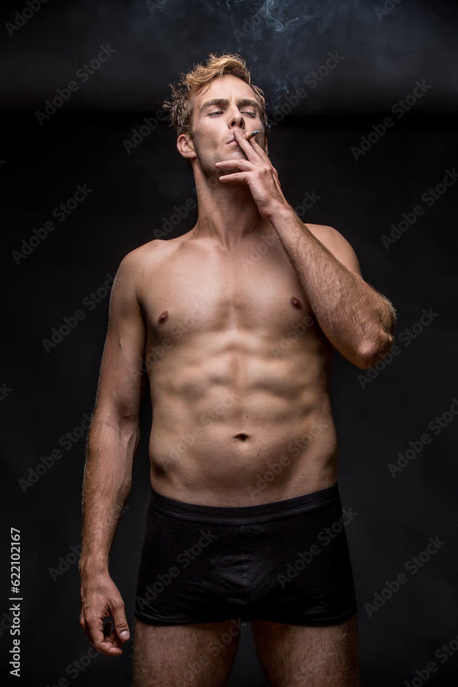 Fototapeta premium Young man with muscular body in black underpants stands on the black background in the studio. He smoke a cigarette and holds it with left hand. Vertical low-key photo.