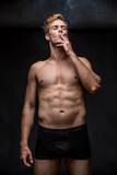 Young man with muscular body in black underpants stands on the black background in the studio. He smoke a cigarette and holds it with left hand. Vertical low-key photo.