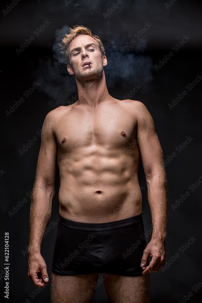 Macho with muscular body in black underpants stands on the black background in the studio. He smoke a cigarette and a smoke swirls around him. Vertical low-key photo. Macho with muscular body in black underpants stands on the black background in the studio. He smoke a cigarette and a smoke swirls around him. Vertical low-key photo.