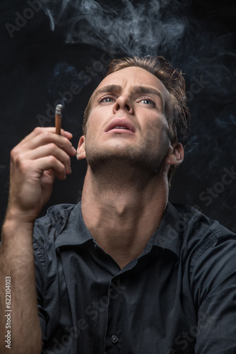 Smoker in dark shirt looks up with parted lips on the black background in the studio. He holds a cigarette in the right hand. Smoke swirls around the man. Vertical low-key photo.