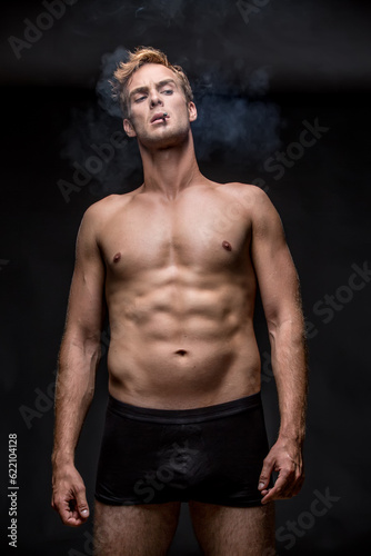 Macho with muscular body in black underpants stands on the black background in the studio. He smoke a cigarette and a smoke swirls around him. Vertical low-key photo.