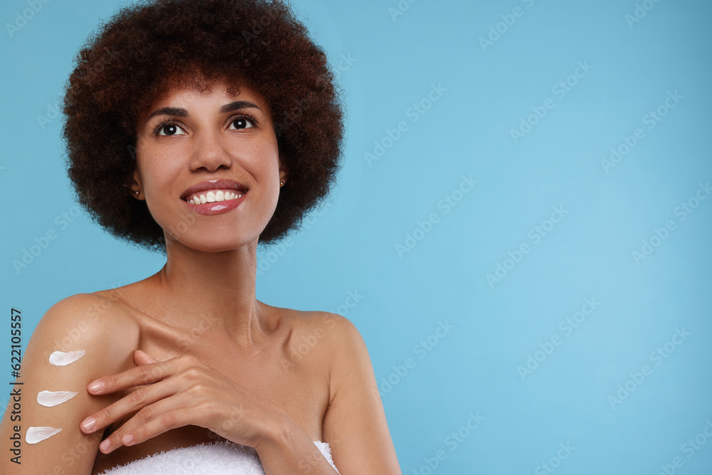 Beautiful young woman applying body cream onto arm on light blue background, space for text