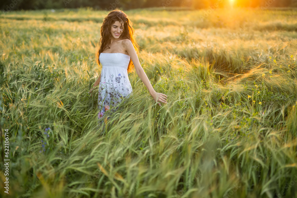 Happy girl stands in the rye field and looks at the left hand on the sunset background. Woman touches the rye. She wears a light dress with prints of flowers. Outdoors. Horizontal.