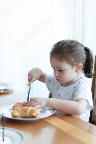 Happy toddler girl eating croissant at the kitchen table, enjoying breakfast with milk or juice. Cozy family home scene, childhood nutrition, cute lifestyle portrait, morning meal.