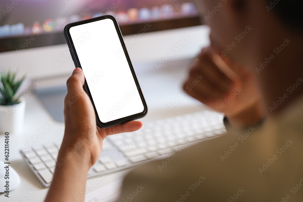 Rear view of young man sitting at his workspace and using smartphone. Blank screen for graphics display montage.