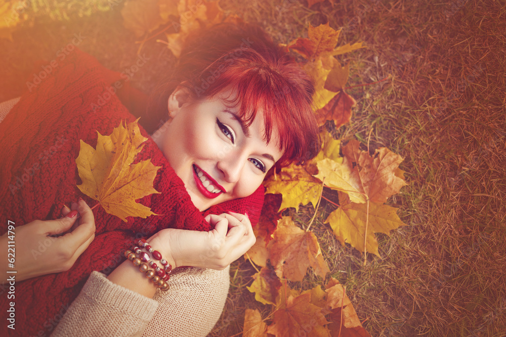 Beautiful young woman with red hair holding yellow maple leaf lying on the ground. Outdoor shot. Fall. Copy space.