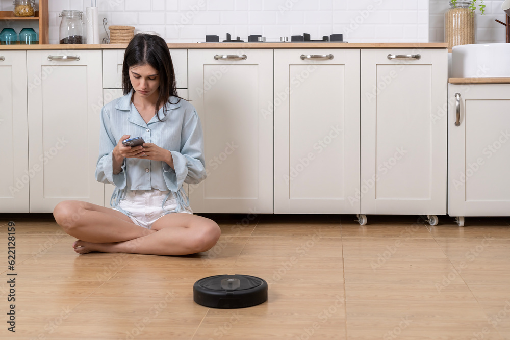 Young beautiful woman sitting on floor with automatic vacuum cleaner in kitchen Young beautiful woman sitting on floor with automatic vacuum cleaner in kitchen