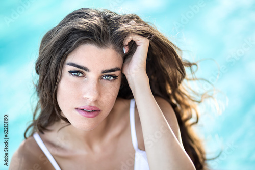Amazing girl in a white swimsuit looks into the camera with parted lips on the background of the swimming pool. She holds her left hand on the head. Outdoors. Horizontal.