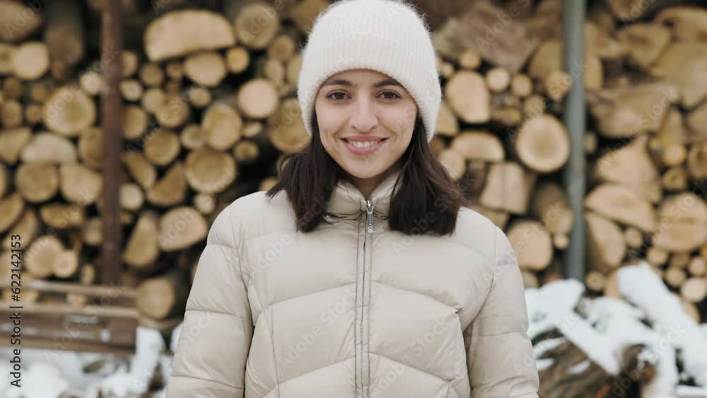 Portrait of a beautiful woman dressed in winter clothes against a background of stacked firewood. The girl smiles at the camera. Snow is falling all around Portrait of a beautiful woman dressed in winter clothes against a background of stacked firewood. The girl smiles at the camera. Snow is falling all around