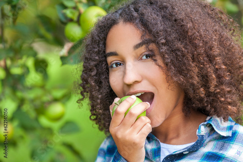 Outdoor portrait of beautiful happy mixed race African American girl teenager female child eating an organic green apple and smiling with perfect teeth photo