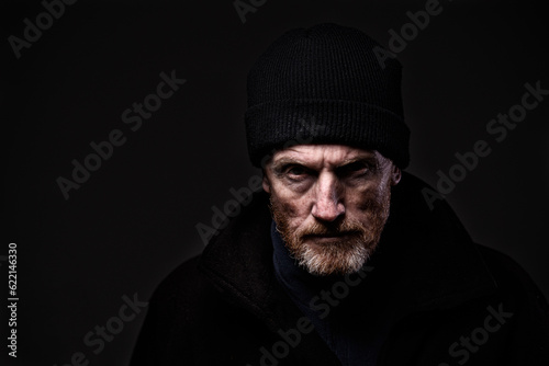 Portrait of a grumpy-looking older, bearded man wearing woollen fisherman's hood, scowling at camera. Dramatic lighting, isolated on black. Ominous, aggressive and dangerous feel. Copy space.
