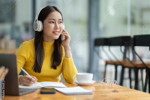 Asian female student or office worker studying online using headphones and laptop taking notes in notebook sitting at coffee shop table relaxing and changing places to work relax.