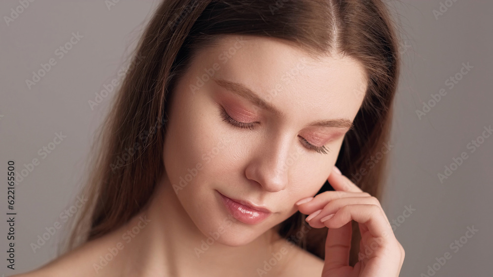 Skin wellness. Beauty treatment. Natural makeup. Young woman with nude eyeshadow and lipstick touching perfect glowing face with hand isolated on beige. Skin wellness. Beauty treatment. Natural makeup. Young woman with nude eyeshadow and lipstick touching perfect glowing face with hand isolated on beige.