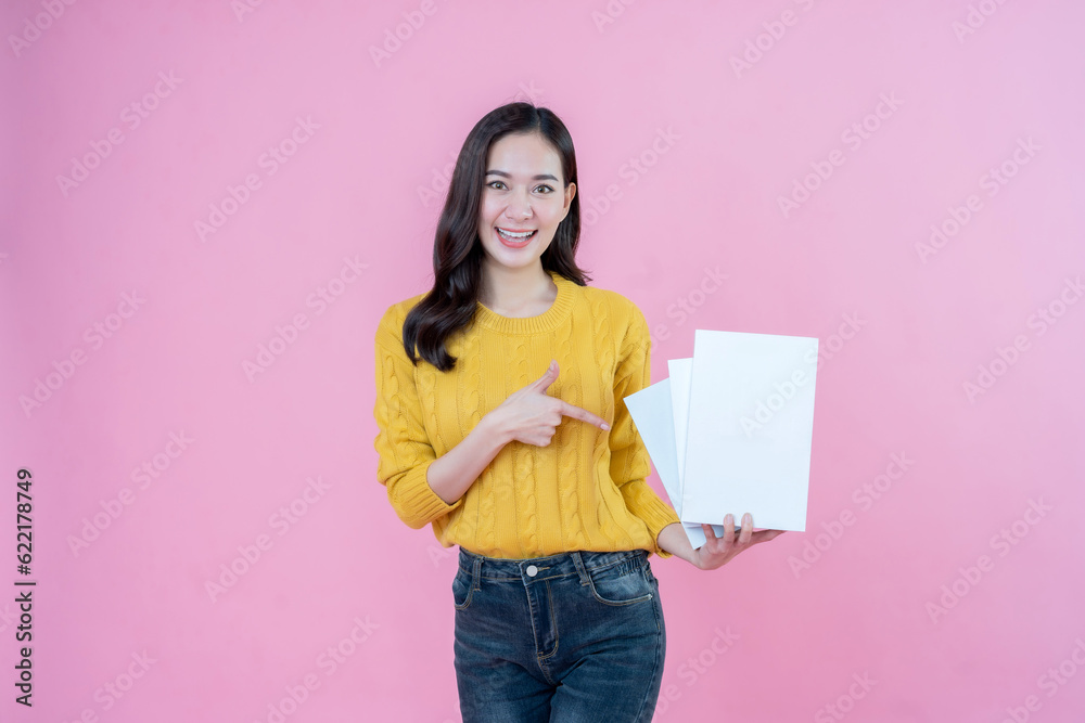 Asian female student with long hair, smiling, pointing to a book in her hand. Presenting about education, the future, wearing a yellow knit coat and jeans. Taking photos in the pink scene studio