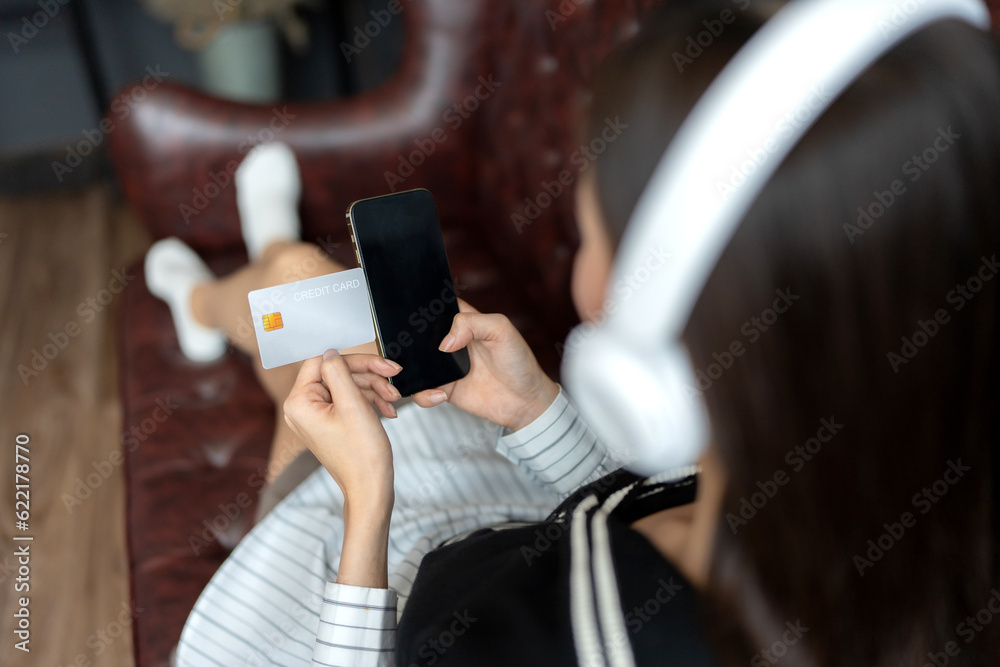 close up of credit card and smartphone In the hands of a long haired asian woman shopping. and check business orders on the sofa wearing wireless headphones in the workplace