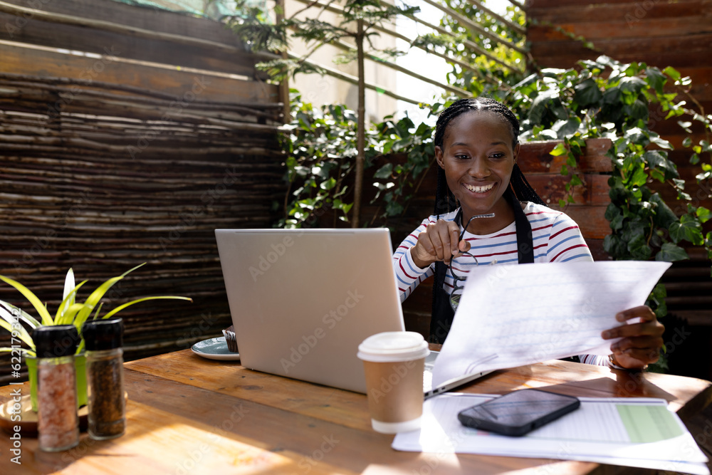 Happy african american female coffee shop owner in apron using laptop at table outside cafe Happy african american female coffee shop owner in apron using laptop at table outside cafe