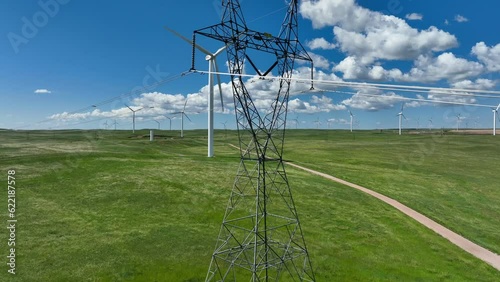High voltage transmission power lines with wind turbine in background. Aerial rising shot in USA plains. Power generation theme. Clean and renewable energy supply near old transmission infrastructure.