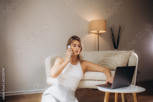 Canvastavla Full length portrait of happy exited woman wearing white shirt and jeans sitting on floor near sofa, using laptop and talking phone