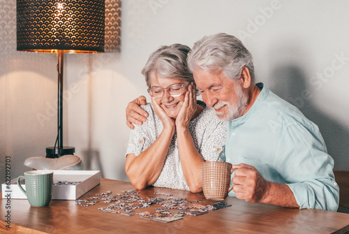 Carefree senior couple at home spend time together doing a puzzle on the wooden table. Elderly man and woman enjoying free time in retirement