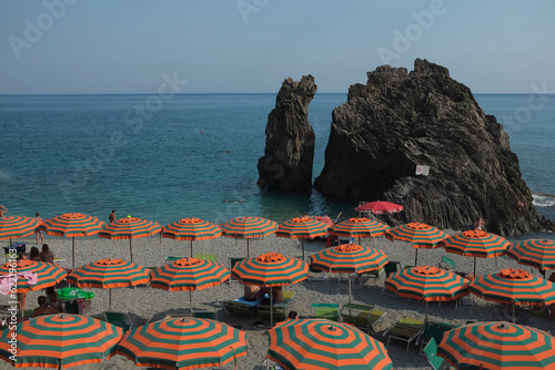 Cinque Terre, Italy - tourists relax under colorful beach parasols in Monterosso al Mare, a seaside village on the rugged Italian Riviera coast. Summer travel vacation background.