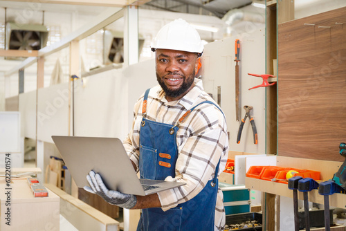 Black male carpenter standing holding laptop looking at camera smiling happy professional hands in furniture carpentry room.